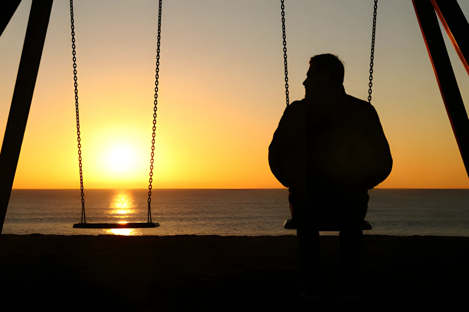 Silhouette of a man sitting alone on a swing at sunset by the sea, with an empty swing beside him